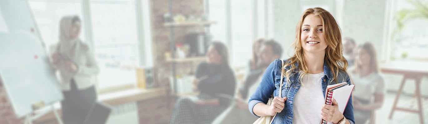 A student standing wearing a shoulder bag and some books in hand, smiling at the camera while a faded picture of a classroom training in background